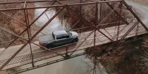 Aerial view of gray pickup crossing narrow rusted truss bridge over river, showcasing truck tires on rural road