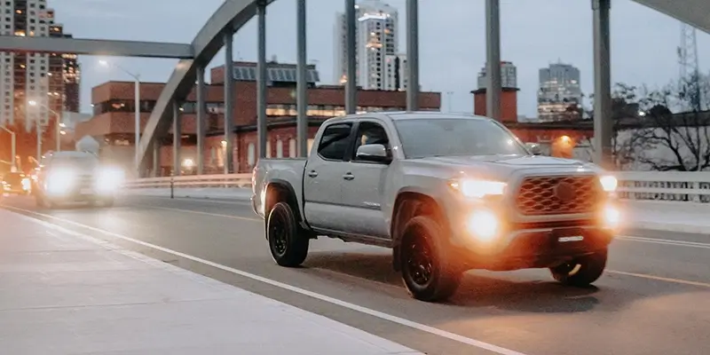 Silver truck with performance tires driving on illuminated urban street at dusk, showcasing city skyline in background.